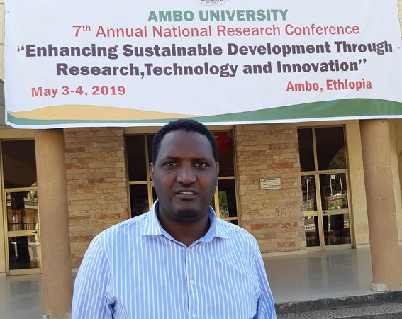 Geda Oncho standing in front of Ambo University, with a big poster in the background