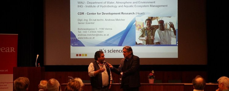 Two people are standing in front of an audience in a lecture hall. One of them is interviewing the other. A large screen in the background shows a presentation. Next to the screen is a red-and-white-coloured roll-up.