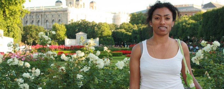 Kasha Tadel Gebre, in a white T-shirt, stands in front of a park with white flowers and a magnificent building in the background. 