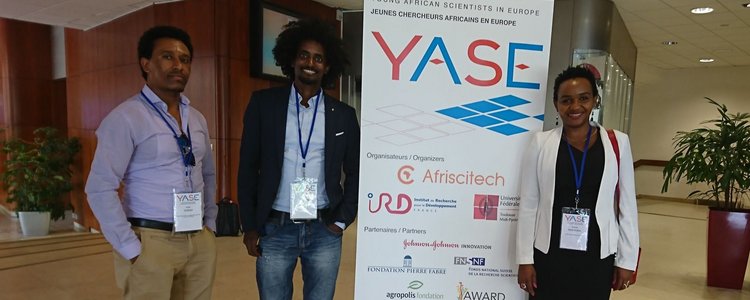 One woman and two men with badges on lanyards around their necks, standing next to a roll-up announcing the "YASE, Young African Scientists in Europe" on 6 July 2018 
