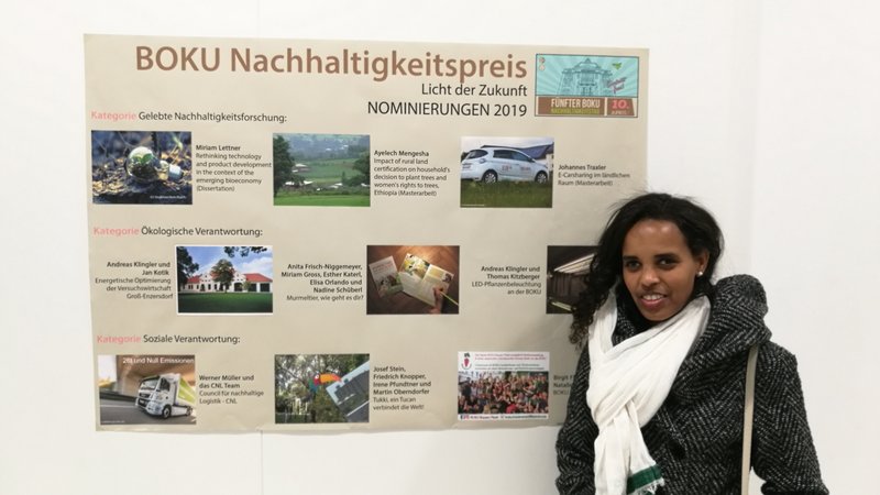 A woman standing in front of a large poster on a wall. The title on the poster reads "BOKU Nachhaltigkeitspreis - Licht der Zukunft - Nominierungen 2019"